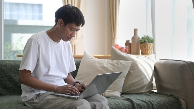 Man using laptop sitting on couch at home chatting or shopping in internet reading message working online with device