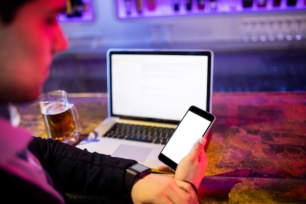 Man using mobile phone with glass of beer and laptop on table at bar counter