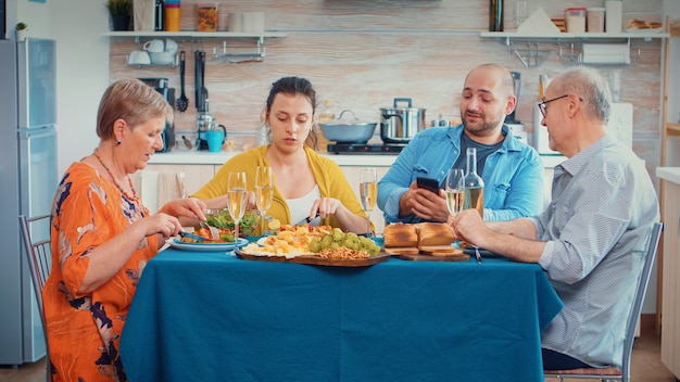 Man using phone during dinner showing some pictures to her\
mother. multi generation, four people, two happy couples talking\
and eating during a gourmet meal, enjoying time at home.
