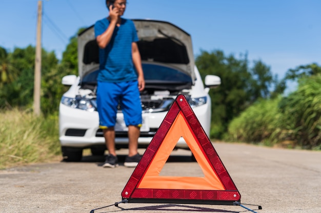 Un uomo che utilizza un telefono mentre ha un problema auto e un segnale di avvertimento triangolo rosso sulla strada