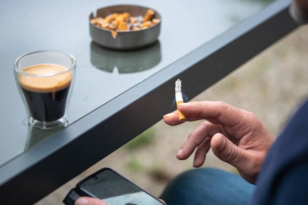 Foto un uomo che usa uno smartphone mentre fuma una sigaretta e ha una tazza di caffè nero accanto a lui