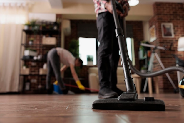 Man vacuuming dirt from the floor while woman wears latex\
gloves and mops. multiracial couple doing housework on day off from\
work, working together to have a harmonious home.