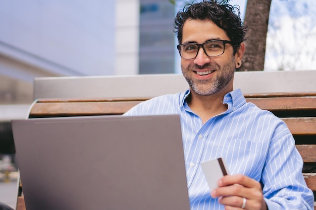 Man van middelbare leeftijd die naar de camera kijkt terwijl hij een creditcard in zijn hand houdt en zijn laptop op schoot