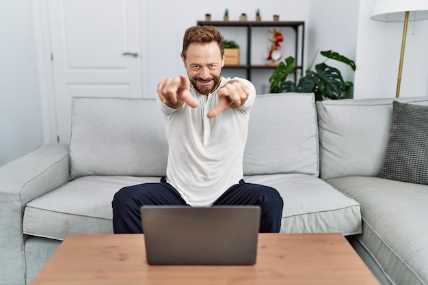 Man van middelbare leeftijd die thuis een laptop gebruikt, wijzend naar jou en de camera met vingers, positief en opgewekt glimlachend