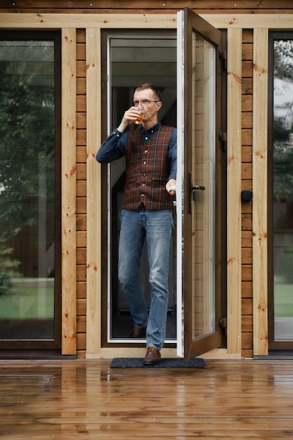 Man van middelbare leeftijd komt uit op het terras van een klein houten huis met een glas in de hand