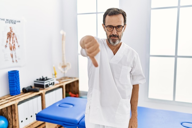 Man van middelbare leeftijd met baard die werkt in een pijnherstelkliniek die er ongelukkig en boos uitziet en afwijzing en negatief toont met duim omlaag gebaar slechte uitdrukking