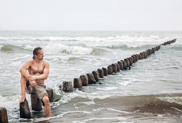 Man van middelbare leeftijd met een naakte torso zit op houten palen op het strand en kijkt in de verte