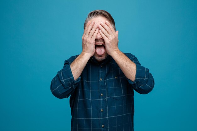 Man van middelbare leeftijd met grijs haar in donkere kleur shirt blij en opgewonden gezicht bedekken met handen uitsteken tong staande over blauwe achtergrond