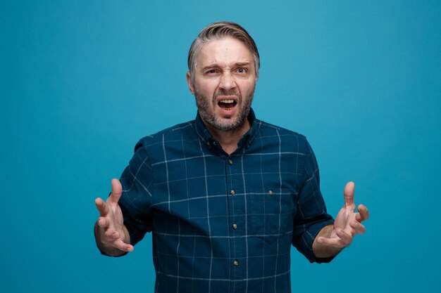 Man van middelbare leeftijd met grijs haar in donkere kleur shirt gek gek schreeuwen met agressieve uitdrukking staande over blauwe achtergrond