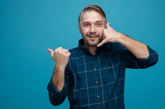 Man van middelbare leeftijd met grijs haar in donkere kleur shirt kijken camera blij en positief tonen bel me gebaar wijzend met duim naar de zijkant glimlachend staande over blauwe achtergrond