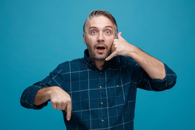 Man van middelbare leeftijd met grijs haar in donkere kleur shirt kijken camera verbaasd maken bel me gebaar met hand wijzend met wijsvinger naar beneden permanent over blauwe achtergrond