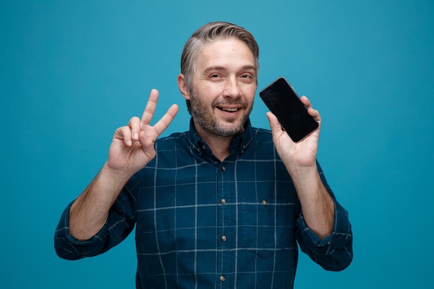 man van middelbare leeftijd met grijs haar in donkere kleur shirt met smartphone met vsign blij en positief kijkend naar camera glimlachend staande over blauwe achtergrond