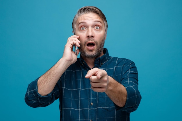 Man van middelbare leeftijd met grijs haar in donkere kleur shirt praten op mobiele telefoon wijzend met wijsvinger naar camera kijken verbaasd en verrast over blauwe achtergrond