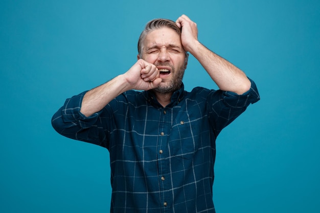 Man van middelbare leeftijd met grijs haar in een donker gekleurd hemd die verward en teleurgesteld bijtende vuist over blauwe achtergrond kijkt