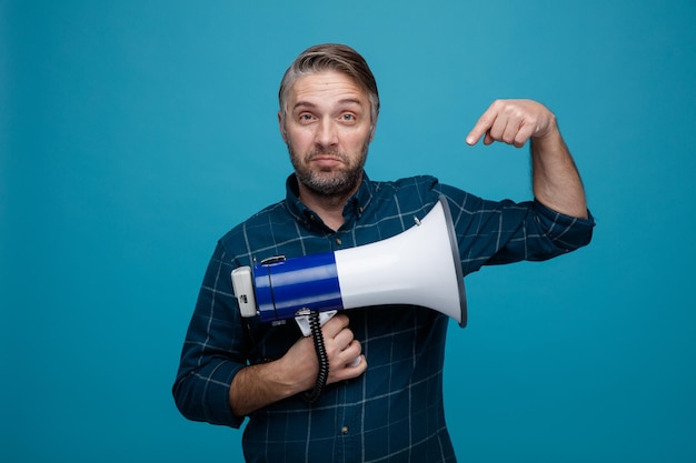 Man van middelbare leeftijd met grijs haar in een donker gekleurd hemd met een megafoon die met de wijsvinger naar hem wijst en glimlachend over een blauwe achtergrond staat