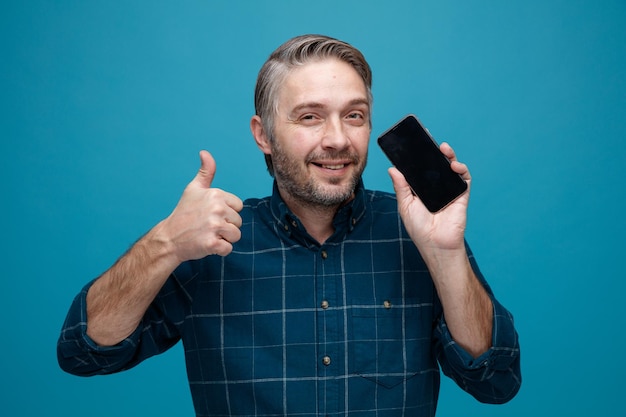 Man van middelbare leeftijd met grijs haar in een donker gekleurd hemd met smartphone die naar een camera kijkt die lacht en duim toont die over een blauwe achtergrond staat