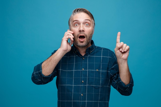 Man van middelbare leeftijd met grijs haar in een donker gekleurd shirt die verrast en verbaasd kijkt terwijl hij op een mobiele telefoon praat met wijsvinger met een nieuw idee over een blauwe achtergrond