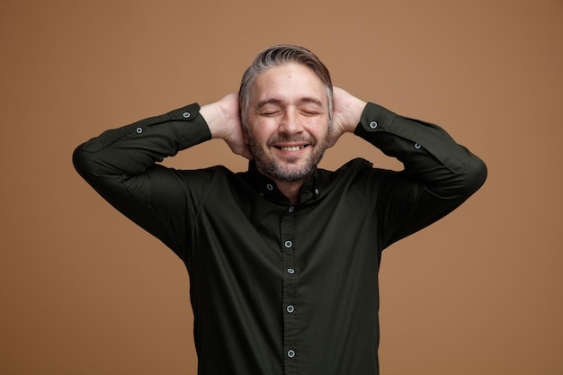 Man van middelbare leeftijd met grijs haar in een donkere kleur shirt blij en opgewonden hand in hand achter zijn hoofd staande over bruine achtergrond
