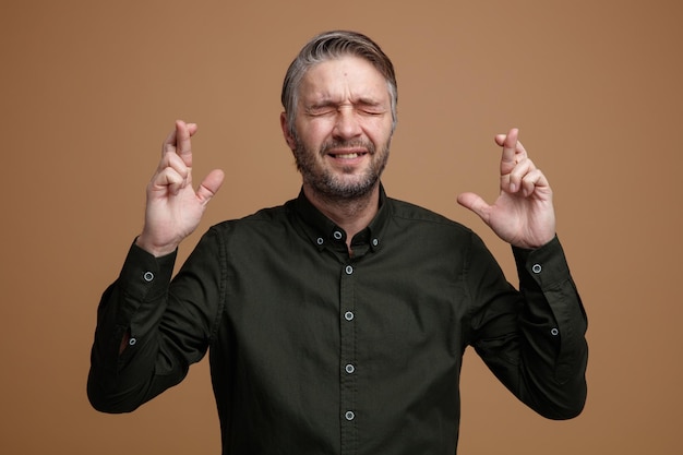 Man van middelbare leeftijd met grijs haar in een donkere kleur shirt die vingers kruist en een wens maakt die opgewonden is over een bruine achtergrond