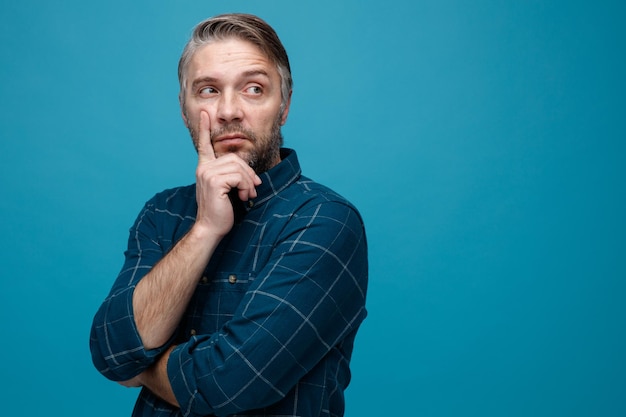 man van middelbare leeftijd met grijs haar in een donkere kleur shirt opzij kijkend met peinzende uitdrukking denkend over een blauwe achtergrond