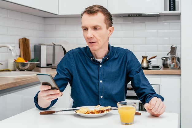 Man van middelbare leeftijd met kort gemberhaar in blauw shirt kijkend naar mobiele telefoon, getroffen door nieuws met opgeheven arm