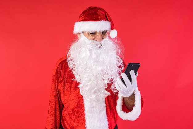 Man verkleed als kerstman die zijn mobiele telefoon raadpleegt op rode achtergrond Kerstviering geschenken consumentisme en geluk concept