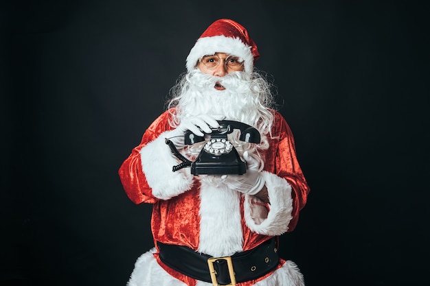 Man verkleed als kerstman met een retro telefoon uit de jaren 60, op zwarte achtergrond. Kerstconcept, Kerstman, geschenken, feest.