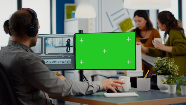 Videografo uomo con auricolare che modifica film in software di post produzione che lavora su schermo verde