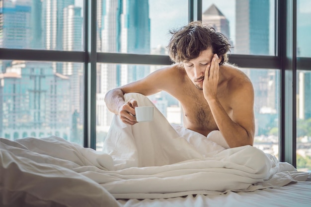 Man wakes up in the morning in an apartment in the downtown area with a view of the skyscrapers and drinks coffee. Life in the noise of the big city concept. Not enough sleep