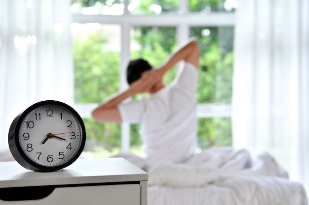 Photo man waking up in the morning sitting on bed and stretching, focus on alarm clock