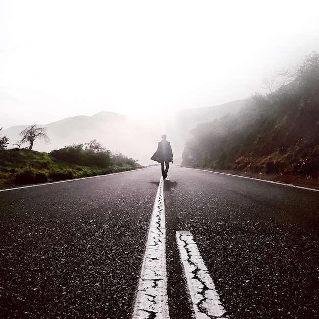 霧の天候で空に向かって山の前で道路を歩いている男