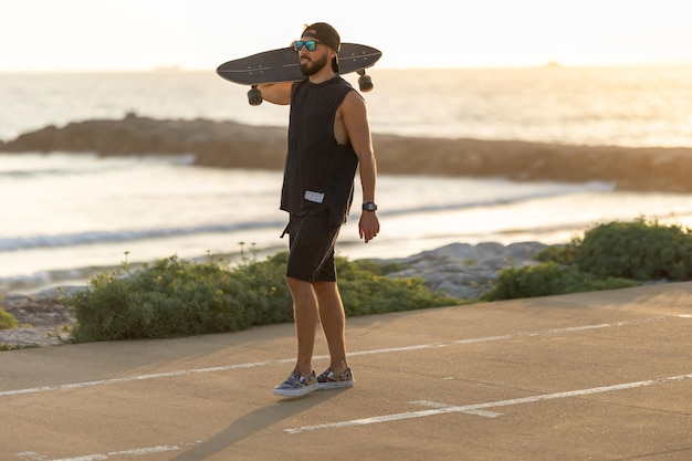 A man walking on the road near by beach holding a skateboard on his shoulder