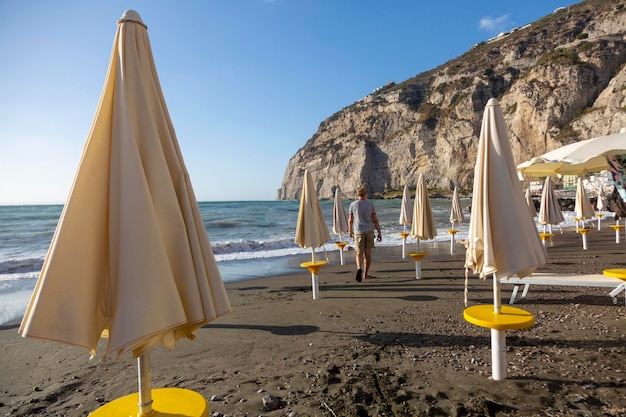 A man walks on a beach with umbrellas and a cliff in the background.