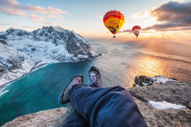 Foto man wandelaar kruis benen zittend op de rotsrug met hete luchtballon vliegen op de oceaan bij zonsondergang