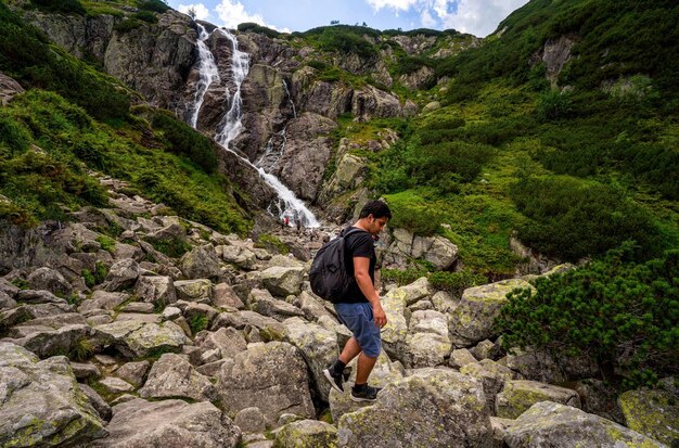 Man wandelaar toerist met rugzak loopt op een rotsachtig berg terrein een enorme waterval in de bergen