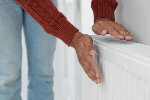 Man warming hands on heating radiator near white wall closeup