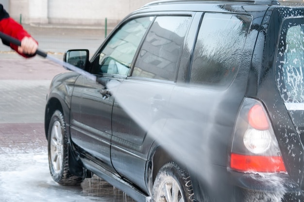 男性がセルフサービスの洗車場で黒い車を強い水で洗います。背面図