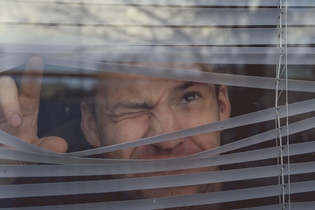 Man watching through window blinds Portrait of young thoughtful male with brown eyes observing through window jalousie