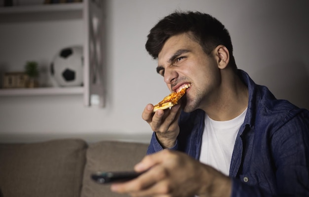 Man watching tv and eating pizza at home at night. Casual guy with remote control and slice of italian food