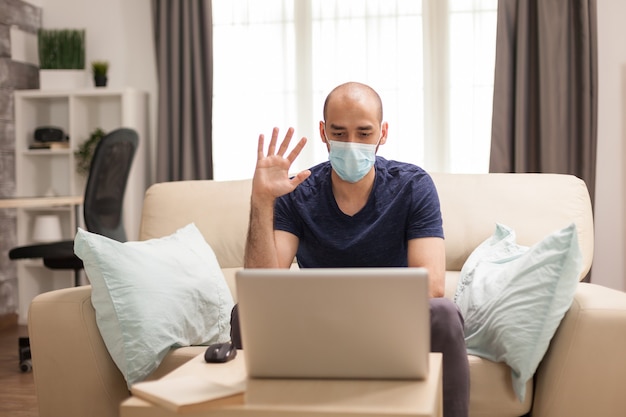 Man waving during a video conference while wearing a protection mask in time of global pandemic.