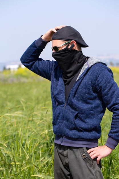 Man wear blue hooded jacket, glasses, cap and mask, took his head and looks at Green Rye. Farmer checking plants on his farm