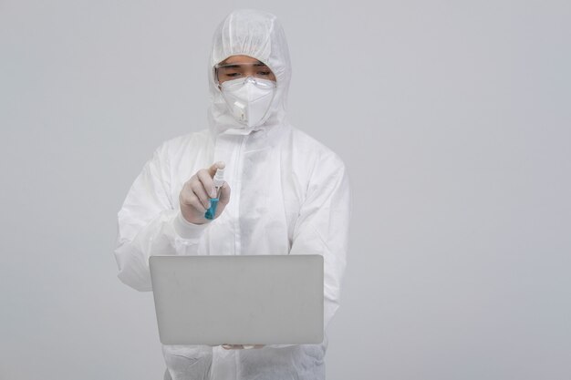 Man wearing biological protective uniform, mask and gloves spraying sanitizer on laptop