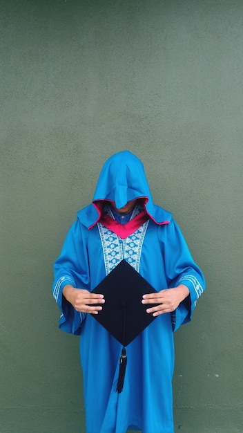 Photo man wearing blue graduation gown holding black mortarboard while standing against wall