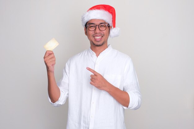 A man wearing christmas hat smiling and pointing to blank card that he hold