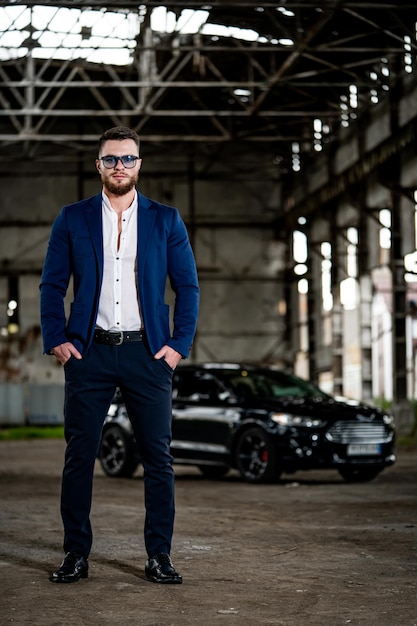 Man wearing dark suit and white shirt inside old broken warehouse Black modern car on the background Front view Full length portrait Closeup