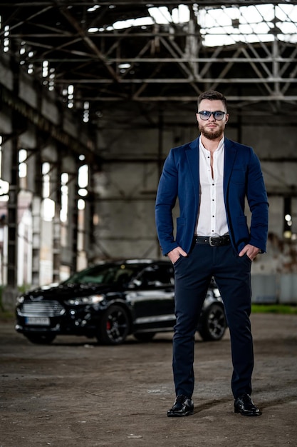 Man wearing dark suit and white shirt inside old broken warehouse. Black modern car on the background. Front view. Full length portrait. Closeup.