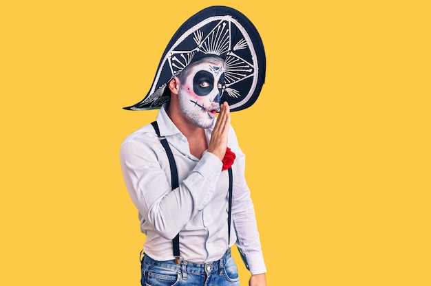 Man wearing day of the dead costume over background hand on mouth telling secret rumor, whispering malicious talk conversation