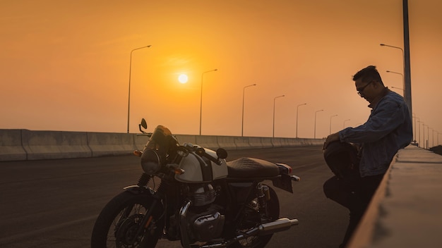 A man wearing a denim jacket, holding a helmet and a vintage motorbike on the highway at sunset. A man riding a vintage motorcycle Biker Lifestyle Concept.