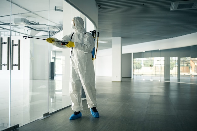 Un uomo che indossa una tuta disinfettante che spruzza con disinfettante le maniglie delle porte di vetro in un centro commerciale vuoto per prevenire la diffusione del covid-19. consapevolezza della salute, pulizia, concetto di difesa.