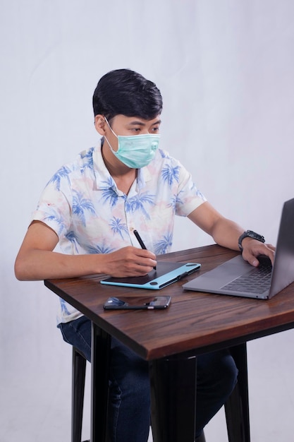 A man wearing a face mask sits at a table with a laptop and a phone.